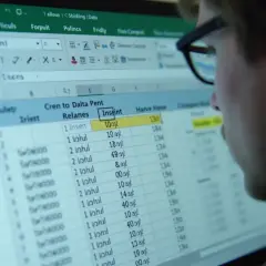 Mężczyzna w okularach analizuje dane w arkuszu kalkulacyjnym Excel. Tabela zawiera kolumny z nazwami i liczbami.