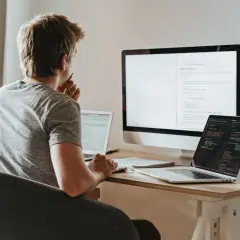 Młody programista pochyla się nad biurkiem, analizując kod na ekranach. Zastanawia się nad optymalizacją zapytań, wykorzystując funkcje agregujące SQL do efektywnego przetwarzania danych.