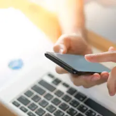 Dłonie trzymają smartfon, na którym widać zawartość w **chrome webview**. Obok leży laptop z wykresami.