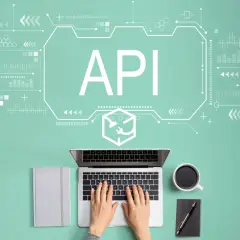 Widok z góry na biurko z laptopem, notatnikiem i kawą. Na ekranie widnieje napis API, symbolizujący interfejs programowania aplikacji.