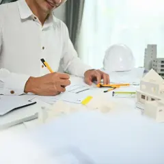 Architekt rysuje plany, obok makieta domu, biały kask i budynki.