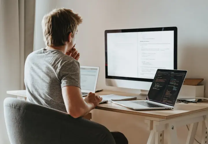 Młody programista pochyla się nad biurkiem, analizując kod na ekranach. Zastanawia się nad optymalizacją zapytań, wykorzystując funkcje agregujące SQL do efektywnego przetwarzania danych.