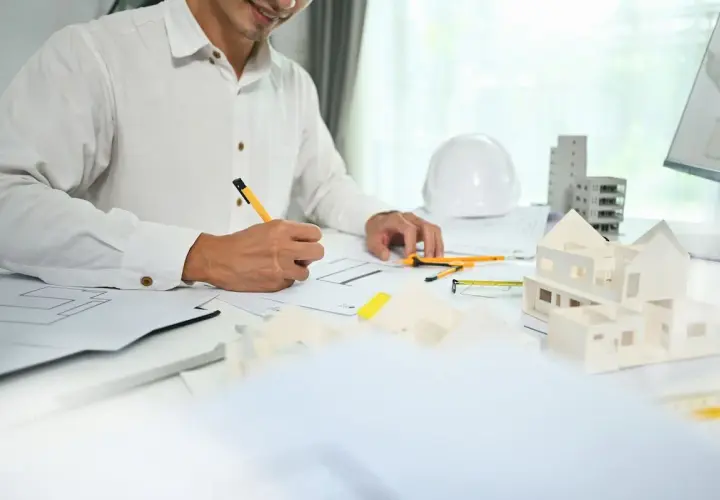 Architekt rysuje plany, obok makieta domu, biały kask i budynki.