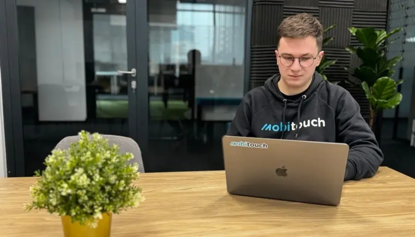 Młody programista w bluzie z logo "mobitouch" pracuje na laptopie. Zastanawia się, co to jest react, tworząc nowoczesne aplikacje.